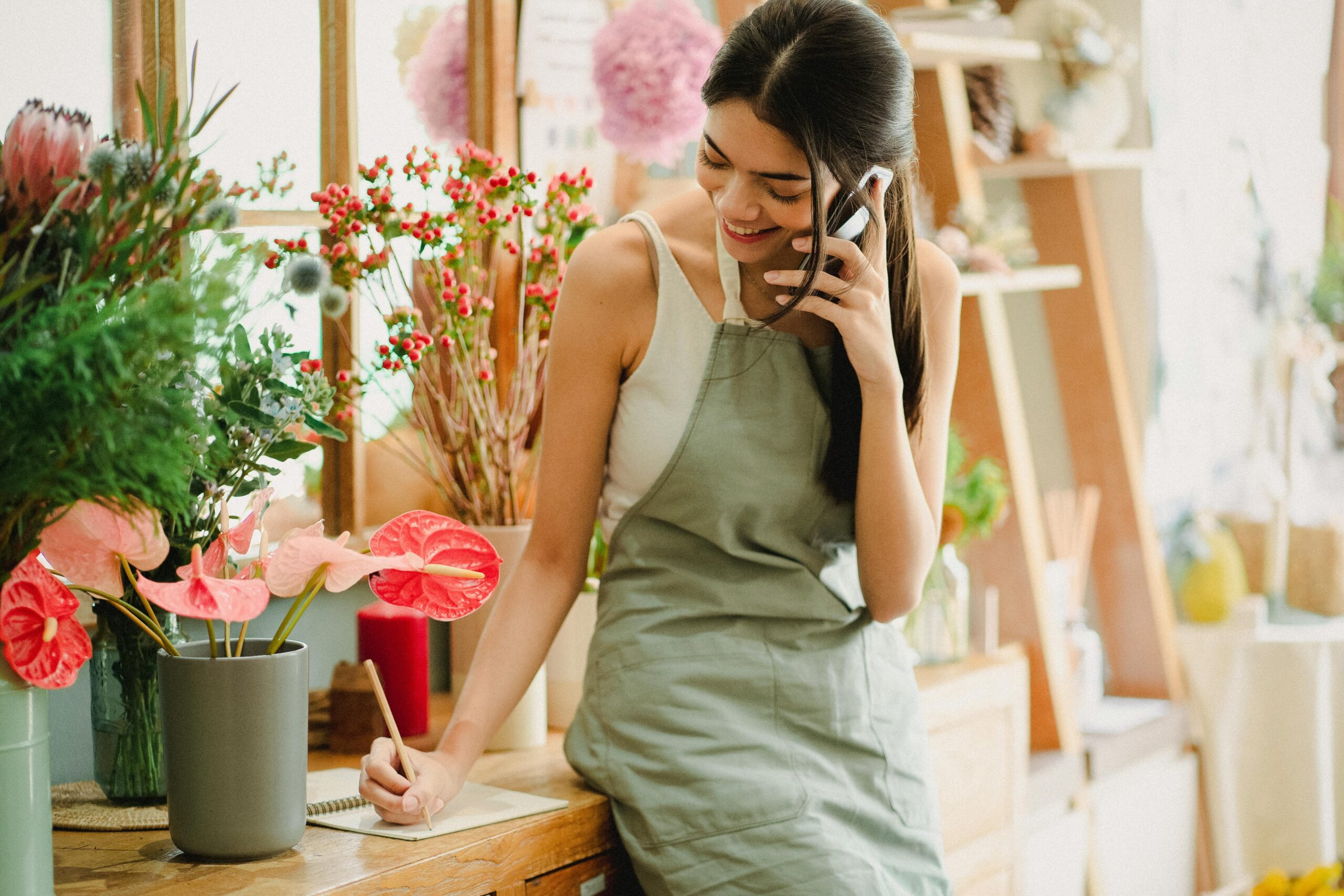 Female florist in apron on phone, writing notes in a floral shop, surrounded by colorful flowers.